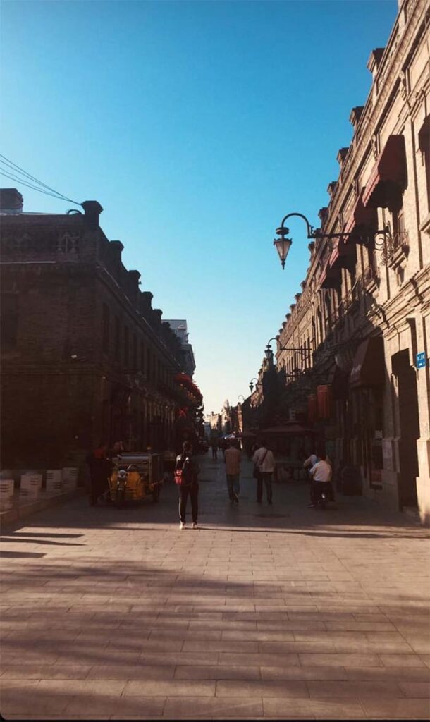 A photo of a street in Harbin, China. The street is not as busy with few people walking. 