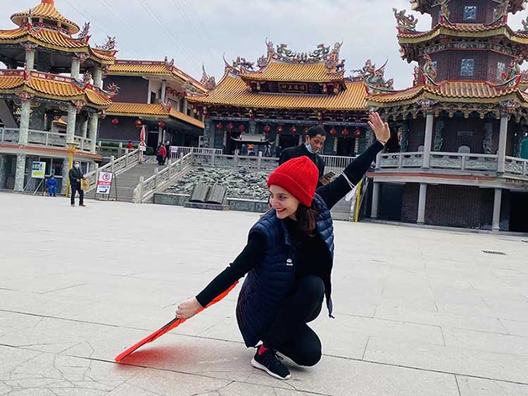 A woman practices taichi with a big red fan on a Square in China. In the back we can see a traditional buddhist temple with orange roofs.