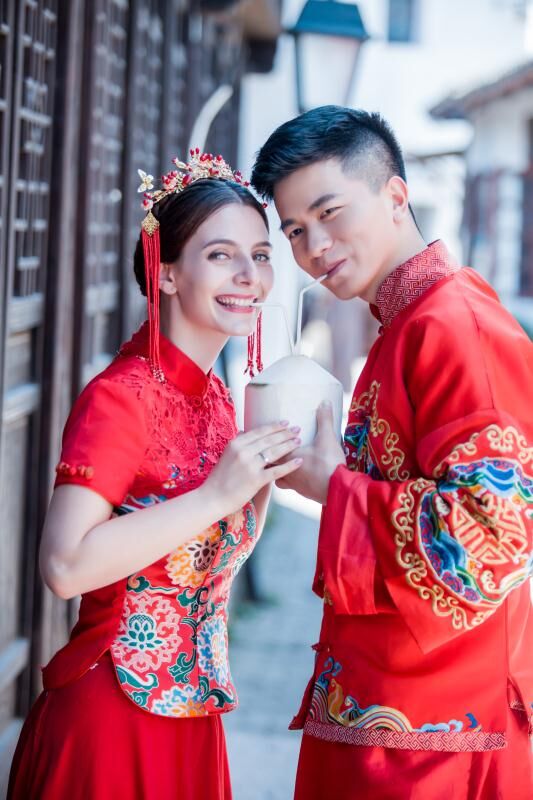 A Chinese man and a French woman drink from a coconut and smile at the camera. They are wearing embroidered red suits, traditional for Chinese weddings.