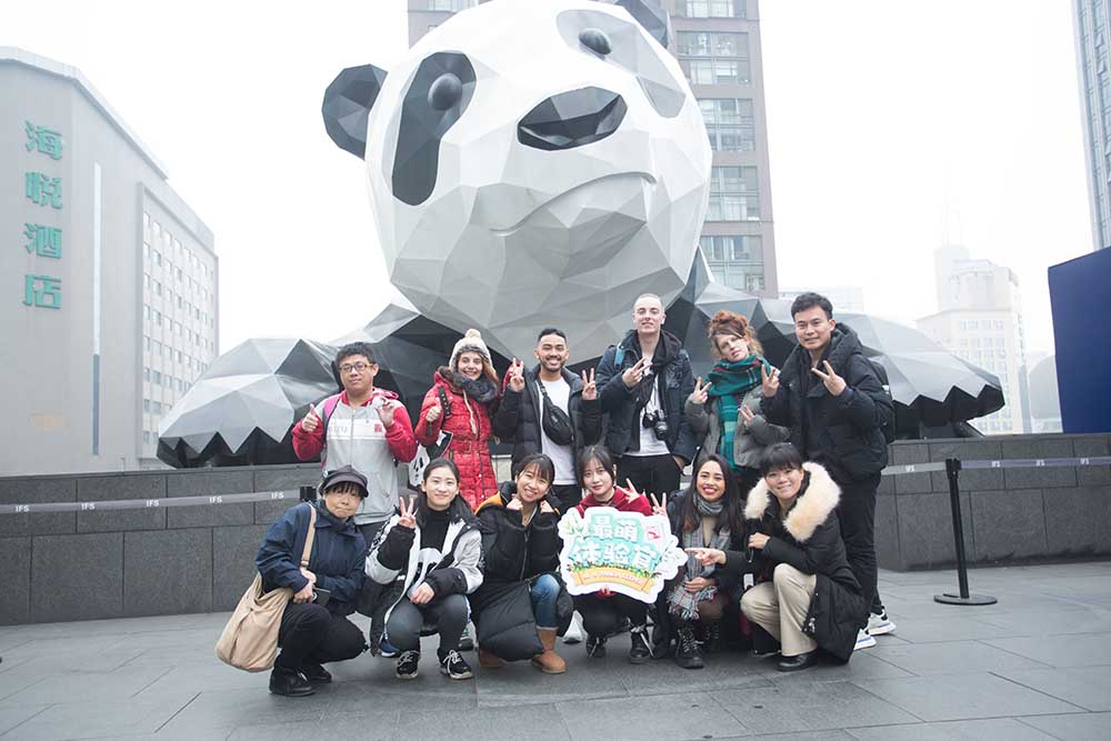 A group of smiley young people pose in front of a Giant Panda sculpture on a winter day, wearing warm clothes, hats and scarves.
