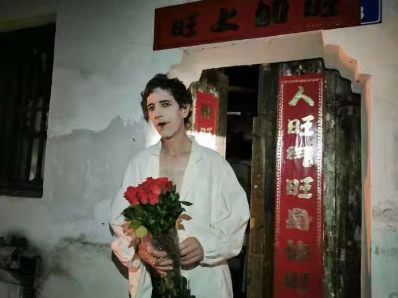 A man wears a wide-open white shirt, white make up on his face with black lips and black make up in his eyes. He holds a bunch of red roses and stands in front of the door of an old house with red and golden Chinese characters on it.
