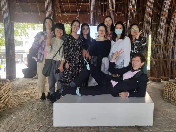 Bill lays down on a white block while eight girls, his students and fellow performers, stand and smile behind him.