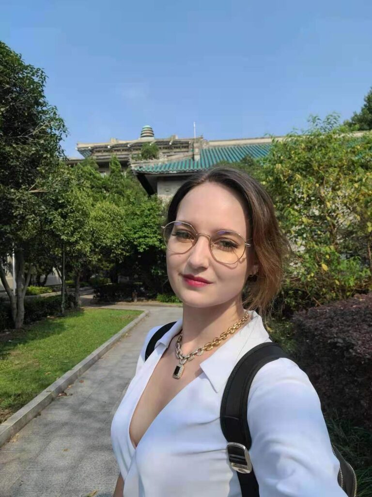 Selfie of a young lady wearing glasses and carrying a backpack in a green garden with green-roofed buildings in the background.