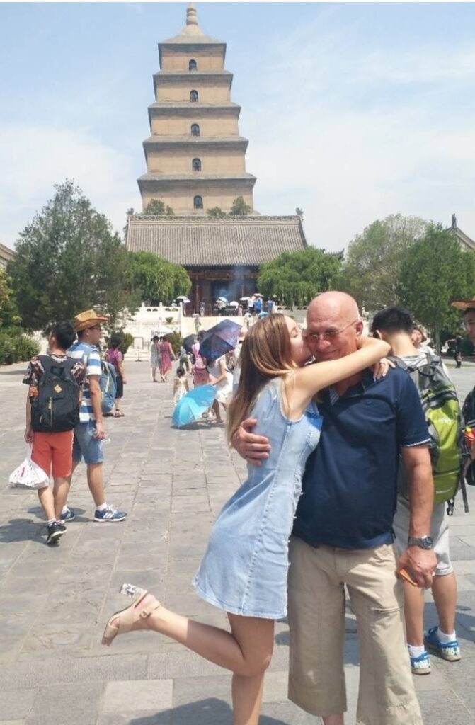 A yound lady hugs and kisses her dad in the cheek. They are standing in front of a crowded square, a traditional Chinese pagoda, the Giant Wild Goos Pagoda in Xi'an is in the background.